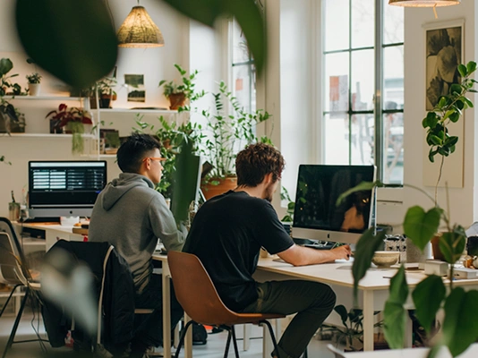 Two men working at computers in a bright office filled with plants and natural light.
