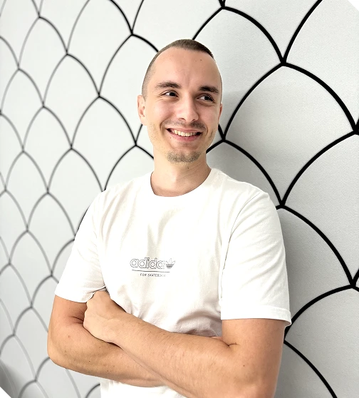 Smiling man with short hair and a light beard wearing a white Adidas t-shirt standing with arms crossed in front of a white wall with black geometric pattern.