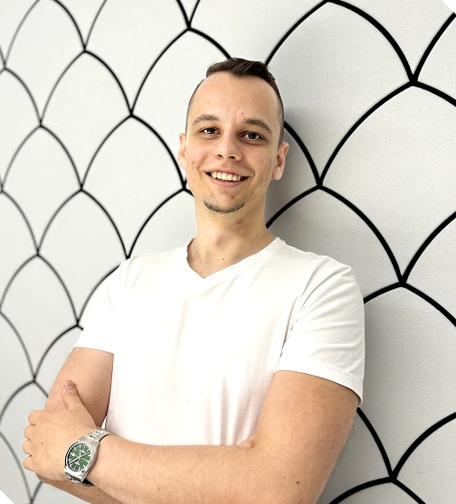 Smiling young man in a white t-shirt standing with arms crossed against a white wall with black geometric patterns.
