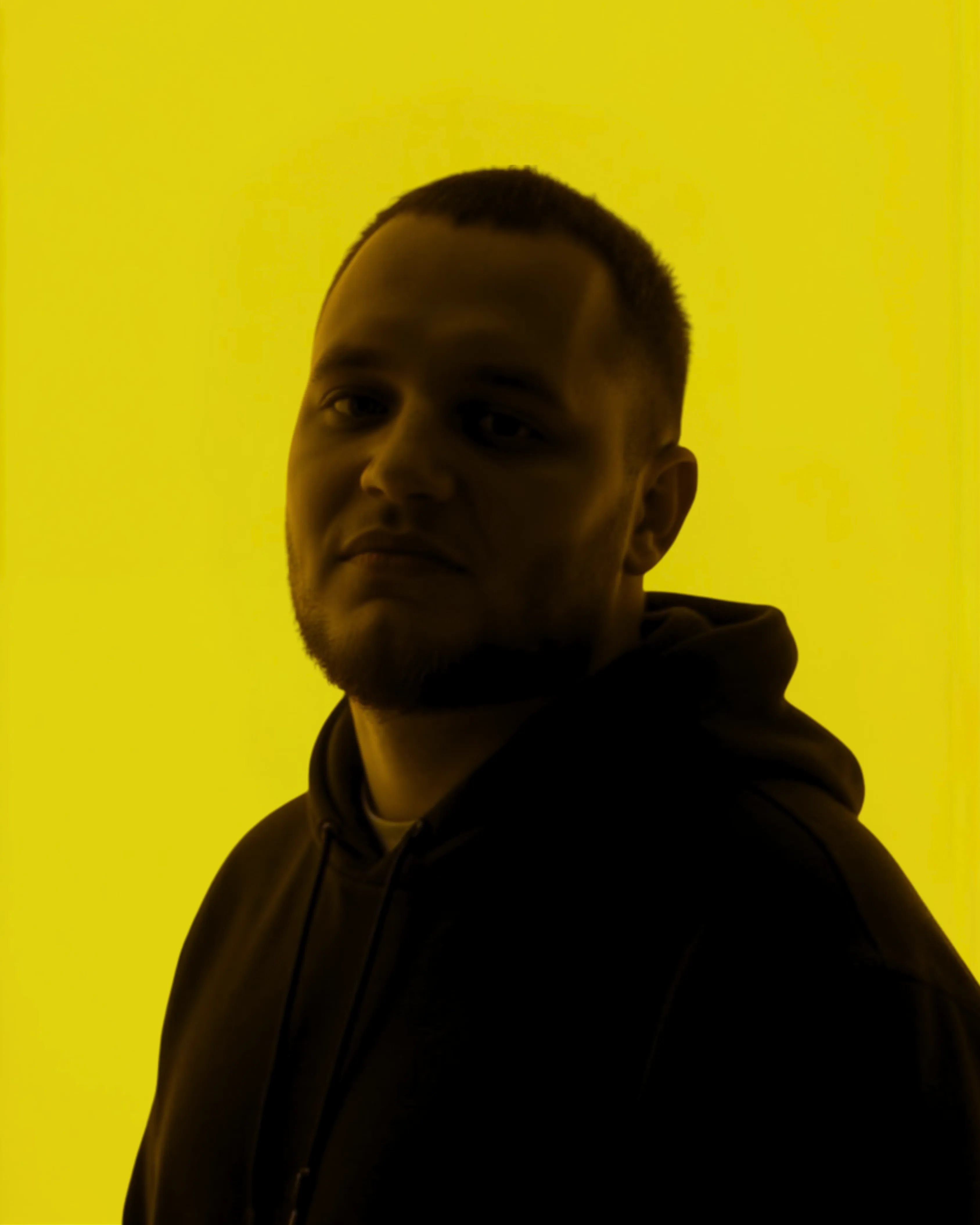 Portrait of a man with short hair and beard wearing a dark hoodie against a bright yellow background.