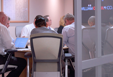 A outside room view of a group of William O'Neil team members having the morning meeting.