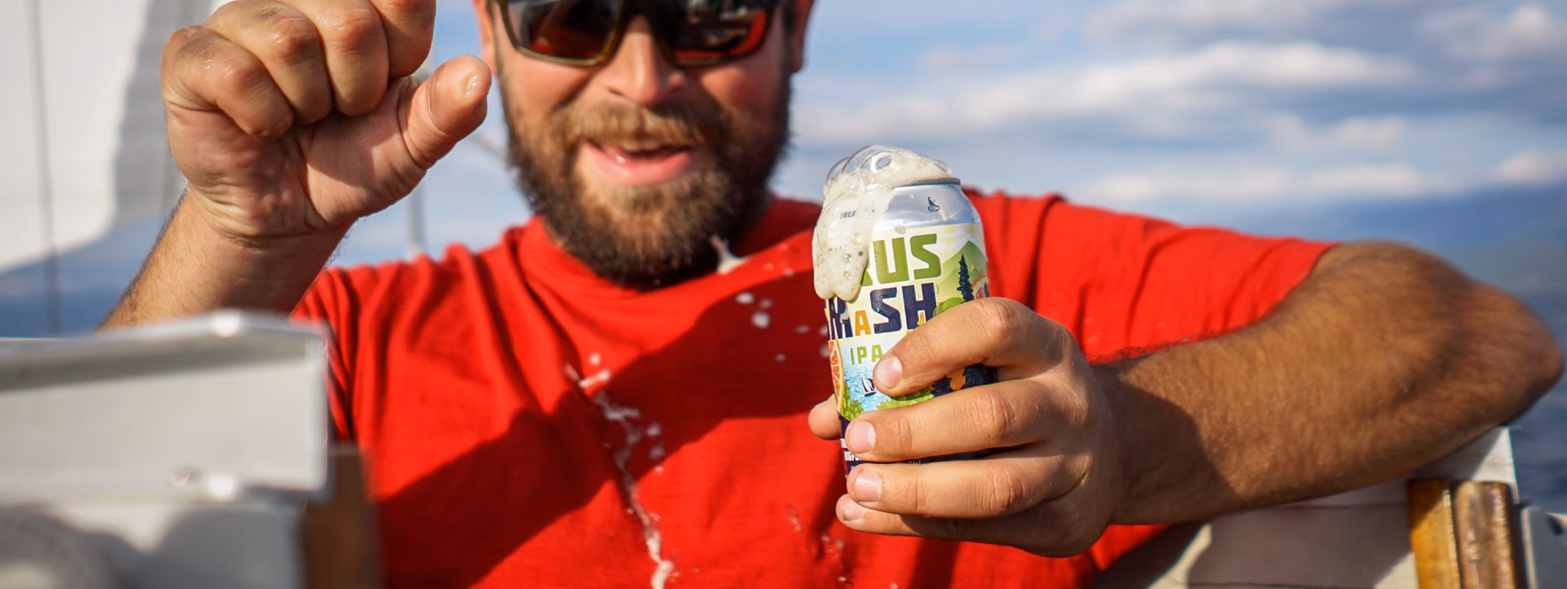 A bearded man in a red shirt smiles widely as he opens a can of fizzy IPA beer, causing foam to spill out.