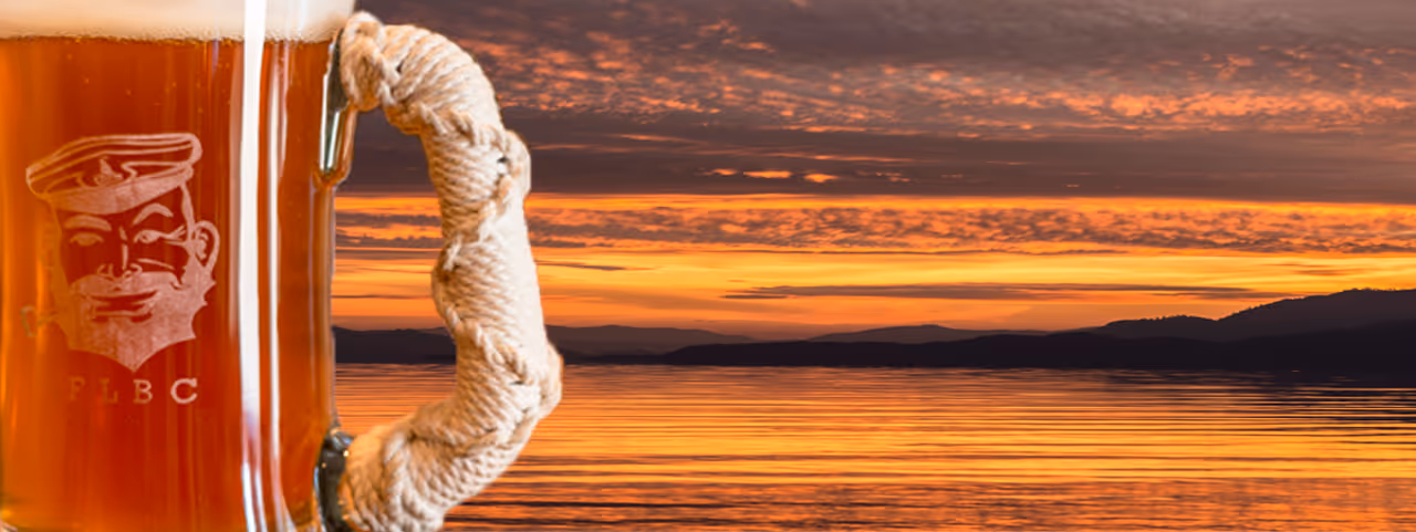 A beer mug with a rope handle is in the foreground, displaying a logo. Behind it, a serene lake reflects a vivid orange sunset, creating a tranquil scene.
