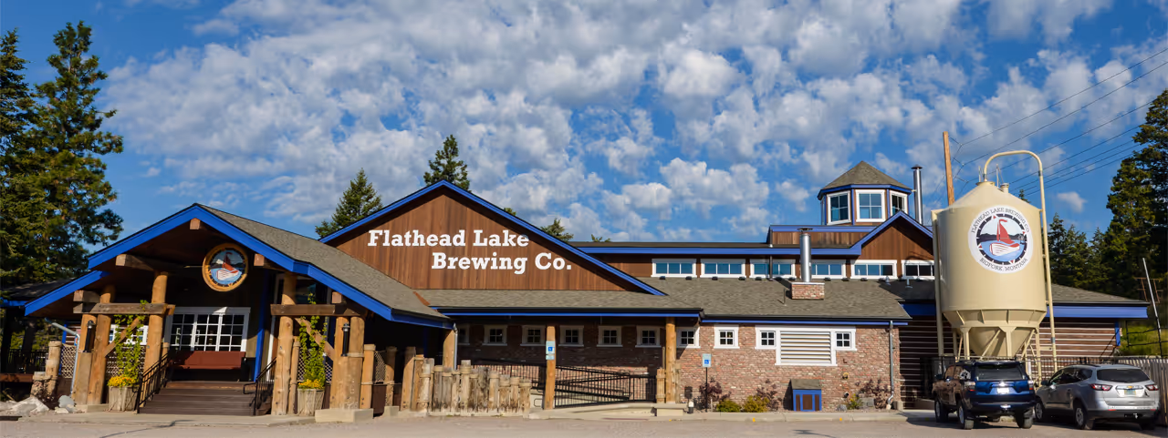 Flathead Lake Brewing Company building with wood accents, set against a blue sky with scattered clouds. Cars are parked nearby.