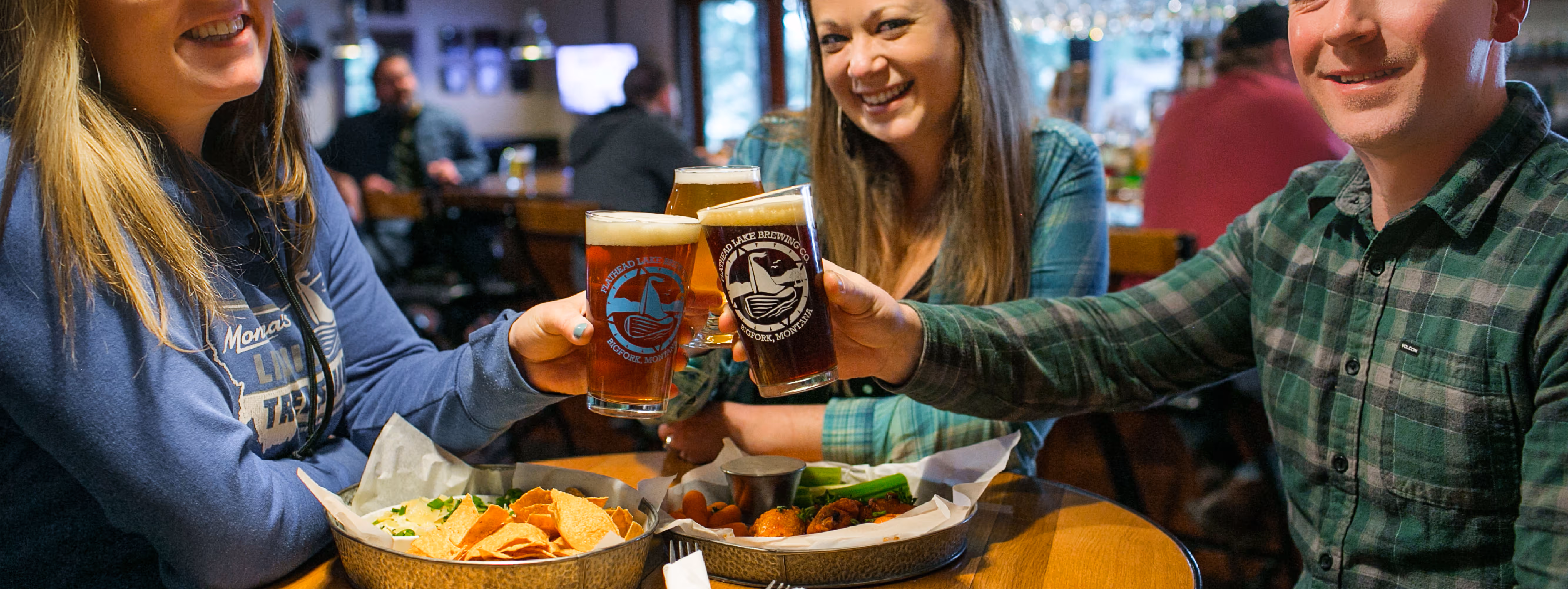Three friends in a pub cheerfully clink glasses of beer over a table with nachos and wings. The scene is lively, warm, and inviting.