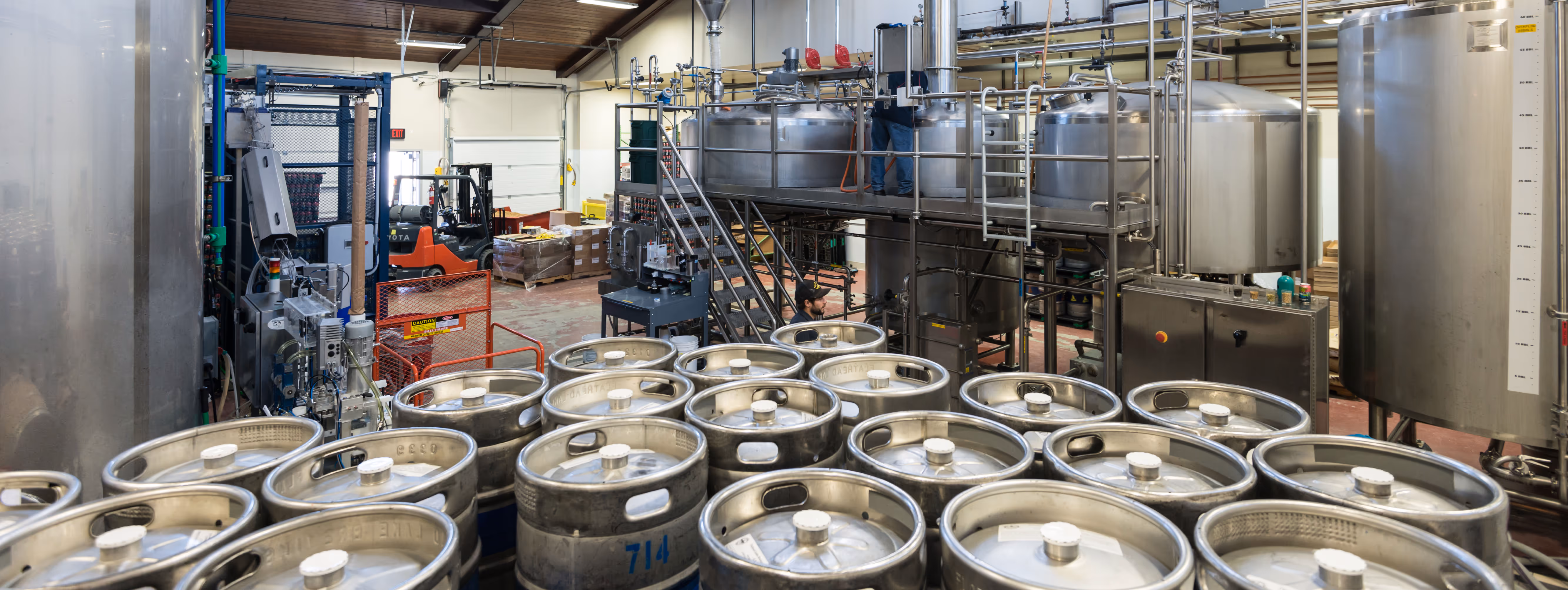 Industrial brewery scene with stainless steel beer kegs in the foreground and large brewing tanks, pipes, and machinery in the background.