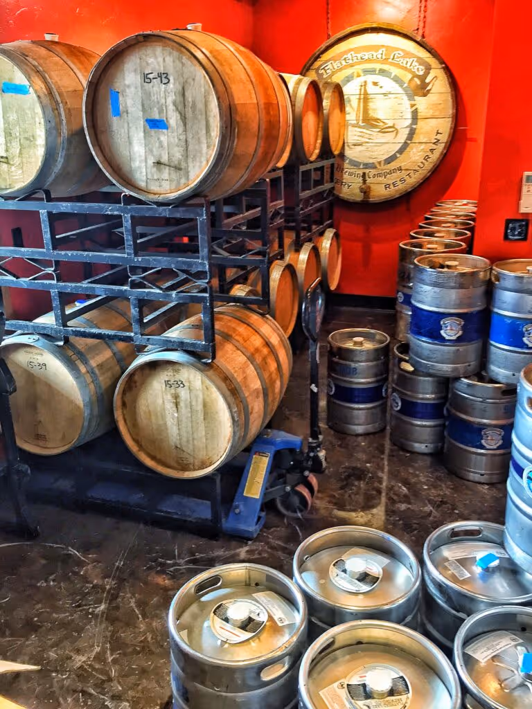 A room with stacked wooden barrels on a metal rack, surrounded by metal kegs.