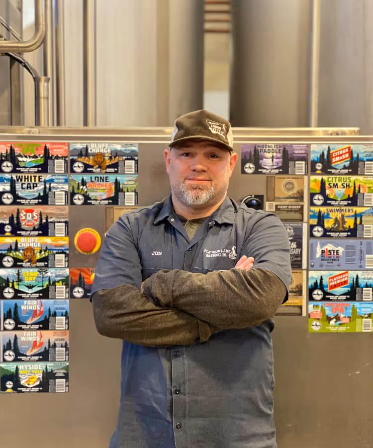 A man in a cap and brewery uniform stands confidently with arms crossed.
