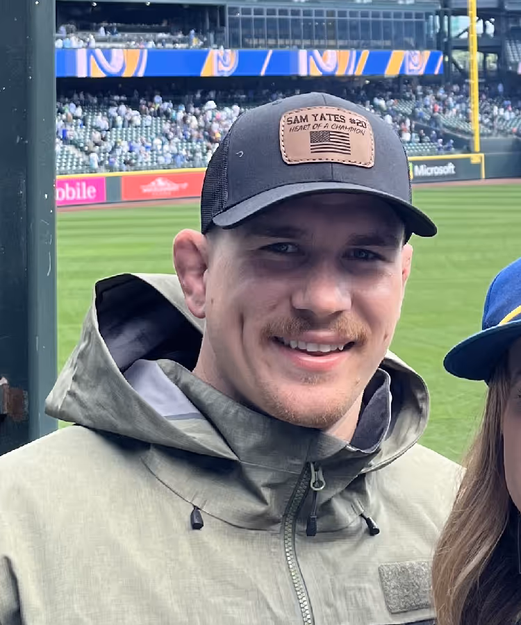 A man smiling at a baseball stadium, wearing a green jacket and a black cap.