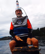 A man in a humorous outfit with a bottle on his helmet sits on a lake.