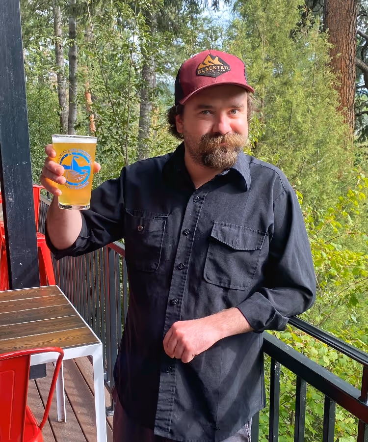 A person in a maroon cap and black shirt smiles, holding a glass of beer on a forested balcony.