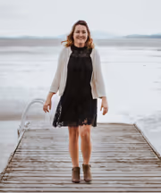A woman in a black dress and light sweater walks on a wooden pier extending over a calm, vast body of water.