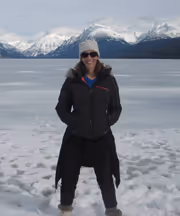 A person in winter clothing stands on snow by a frozen lake, with snow-capped mountains in the background.