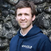 Man with brown hair wearing a navy blue hoodie with 'ENVOLA' logo, standing in front of a stone wall.
