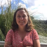 Smiling woman with shoulder-length brown hair wearing a red floral dress, standing outdoors with tall grasses and a partly cloudy sky in the background.