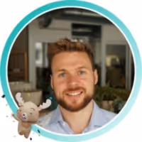 Smiling man with short brown hair and beard wearing a light blue shirt, with a cartoon moose character overlay.