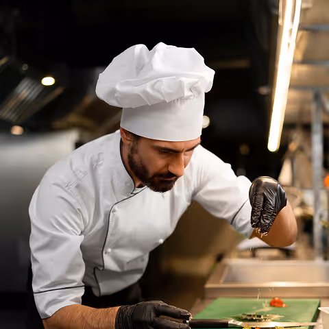 Chef in a white uniform and hat carefully garnishing food with black gloves in a professional kitchen.