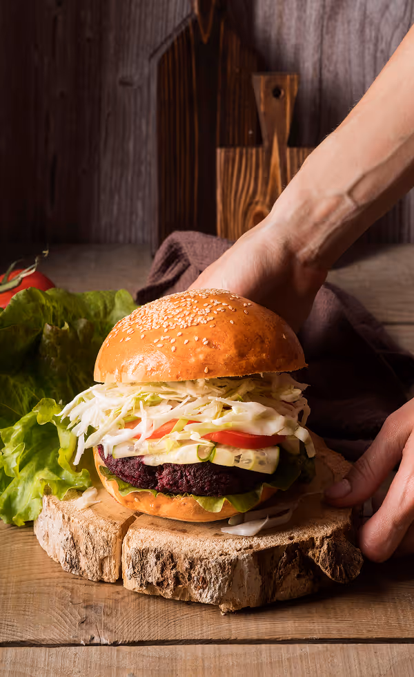 Hand holding a sesame seed bun burger with lettuce, tomato, cabbage, and a beet-based patty on a wooden slice.