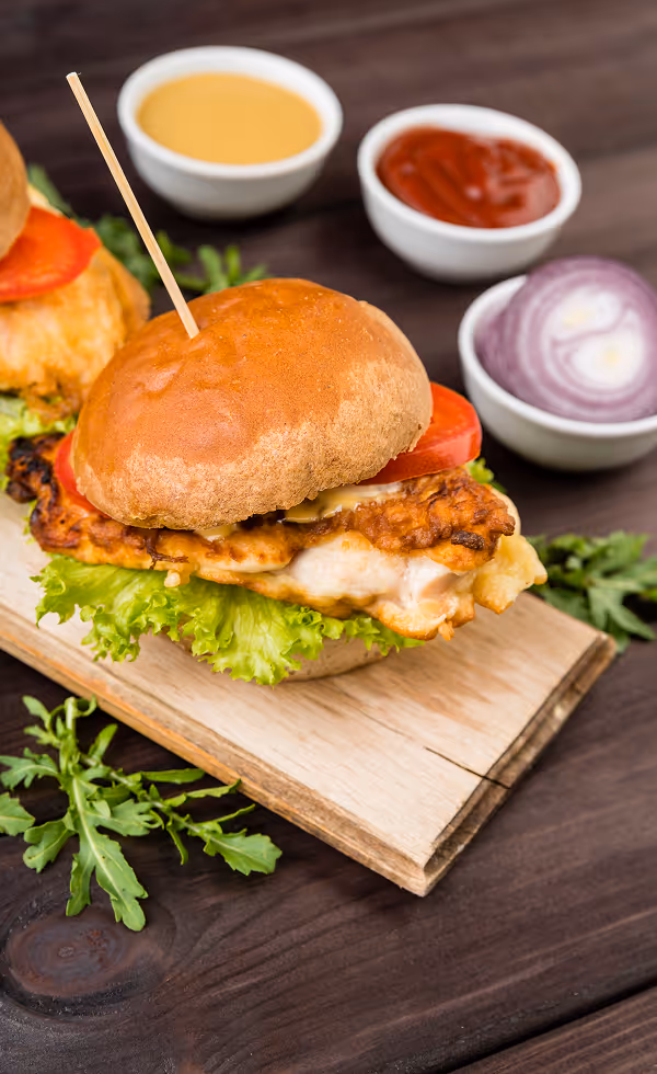 Grilled chicken sandwich with lettuce and tomato on a wooden board, accompanied by bowls of sauce and red onion slices.