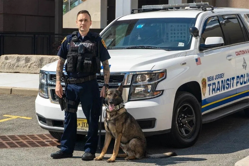 A New Jersey Transit Police K9 officer stands beside his trained police dog in front of a marked patrol vehicle.