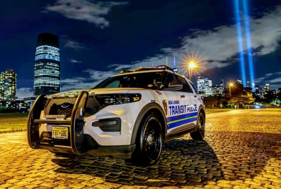 A New Jersey Transit Police SUV parked on a cobblestone street at night with the city skyline and twin beams of light in the background.