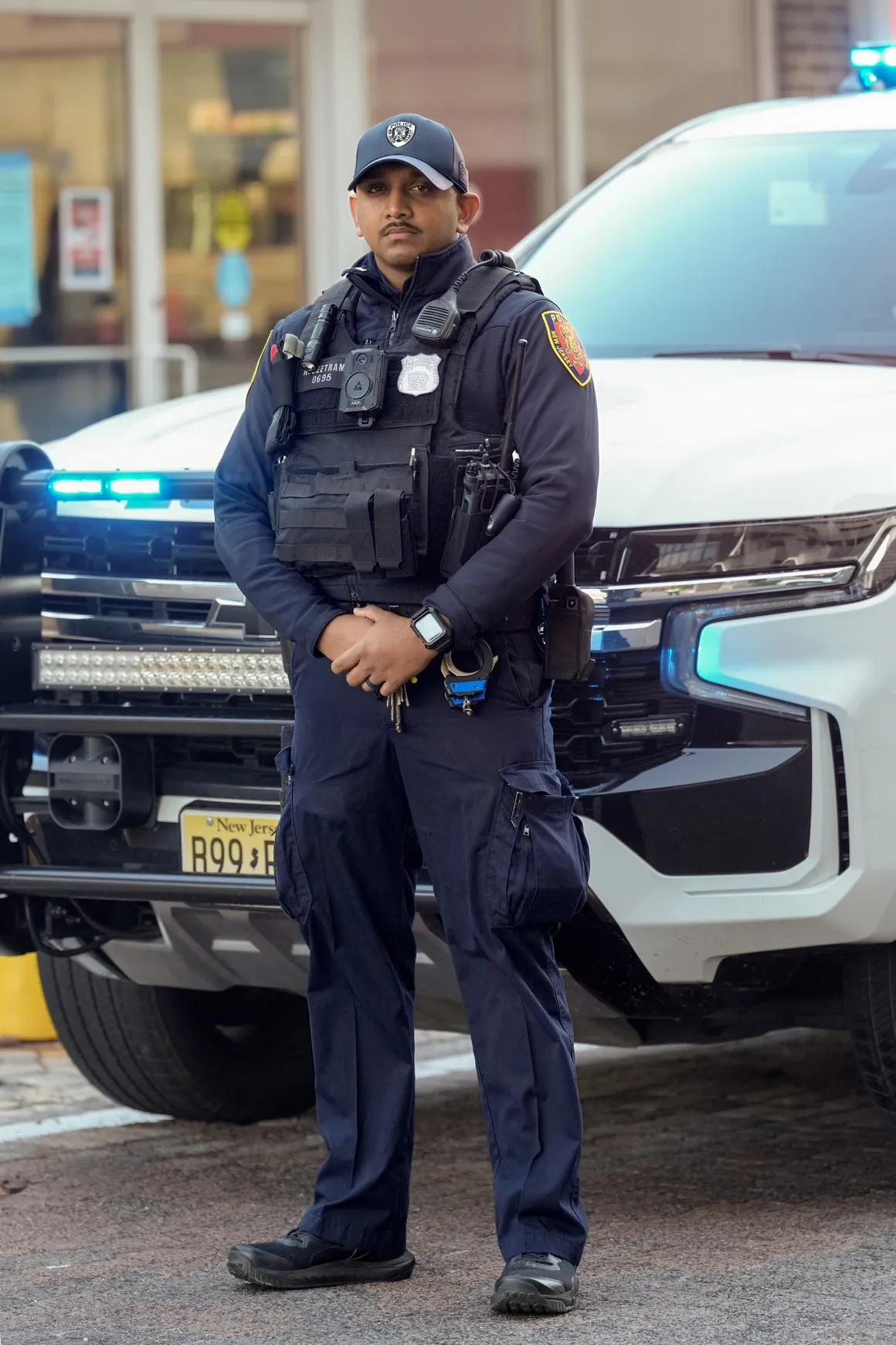 A New Jersey Transit Police officer stands confidently in front of a marked patrol vehicle with flashing lights on a city street.