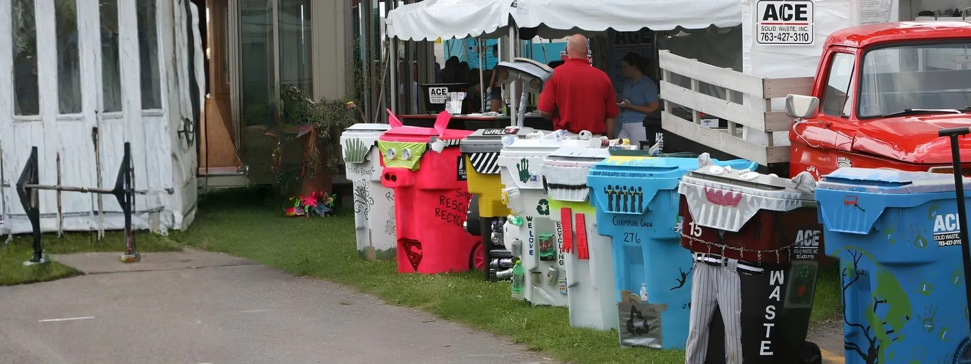 Line of decorated recycling bins on grass near a red truck and white tents at an outdoor event.
