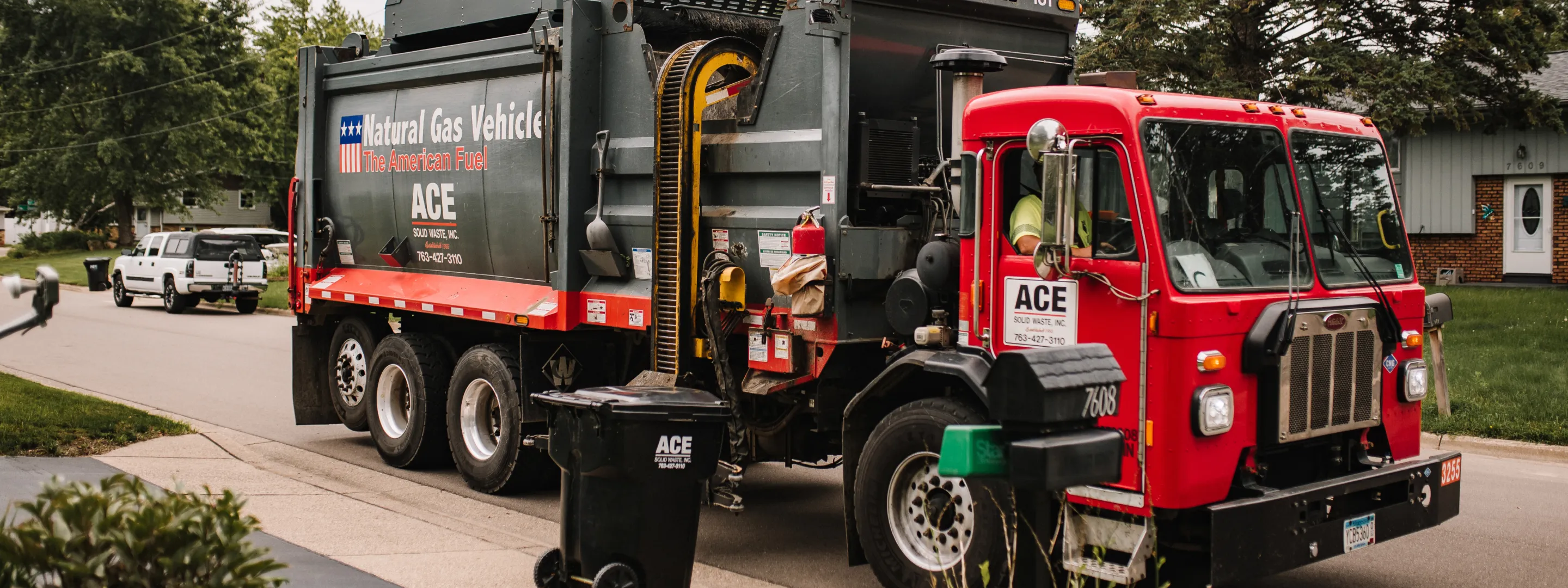 Red and gray ACE Solid Waste natural gas garbage truck on a suburban street next to a trash bin.