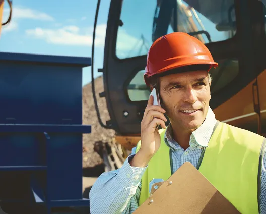 Construction worker in an orange helmet and vest talking on phone, holding clipboard by machinery.