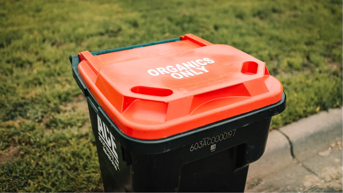 Residential organic waste cart with an orange lid labeled ORGANICS ONLY on grass near a curb.
