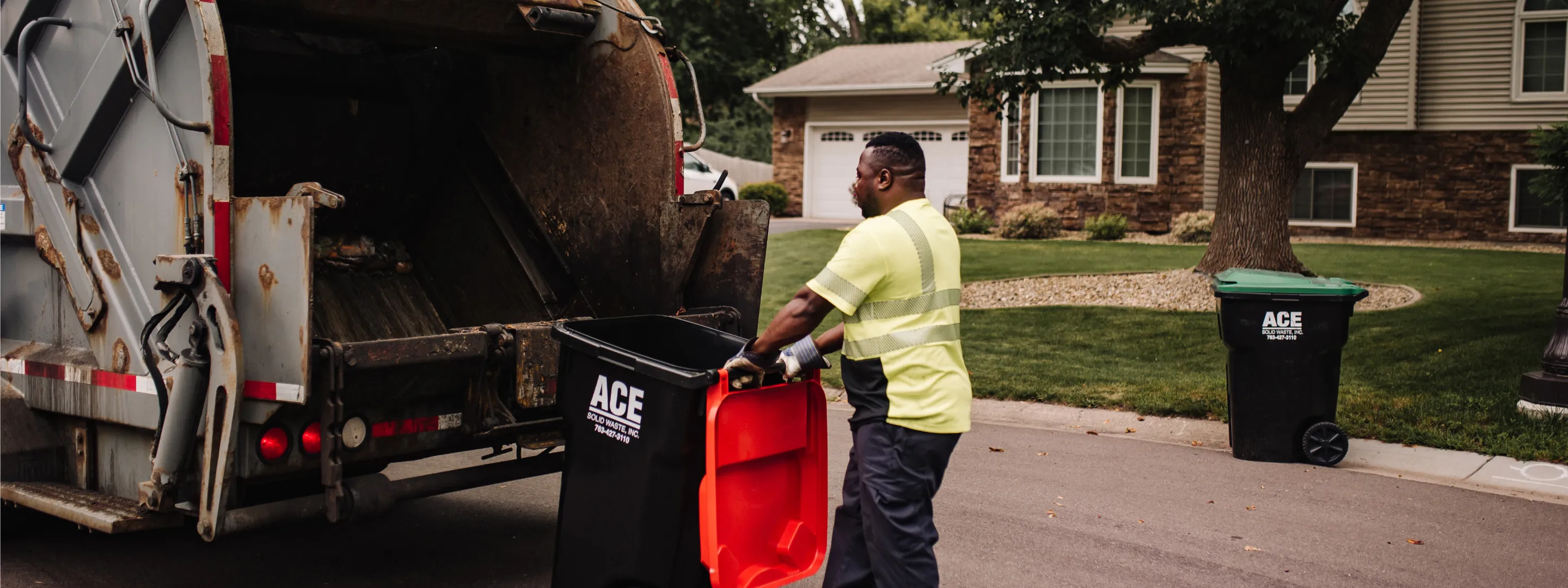 Waste worker in yellow vest loading black and red trash bin into garbage truck on residential street.
