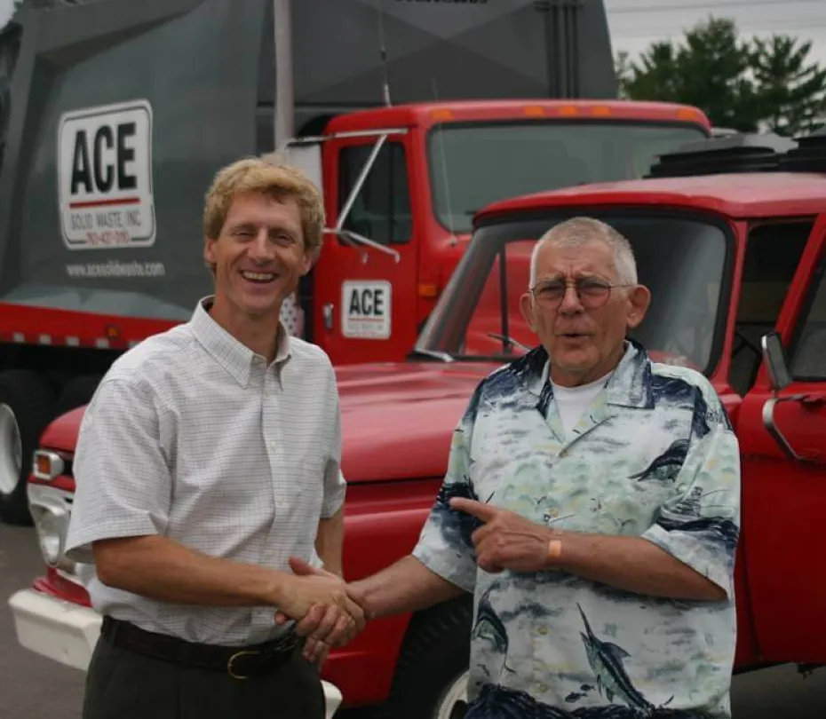 Two men shaking hands in front of red ACE Solid Waste trucks in an outdoor setting.