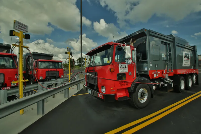 Red Ace Disposal trucks connected to fueling stations under a partly cloudy sky.