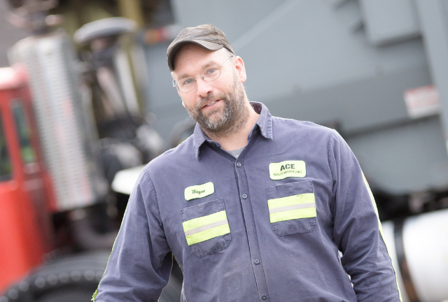 Man in work uniform and cap standing outdoors with truck in the background.