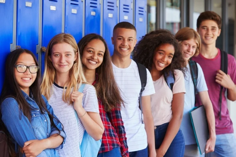Group of diverse smiling teenage students standing in front of blue lockers with backpacks and folders.