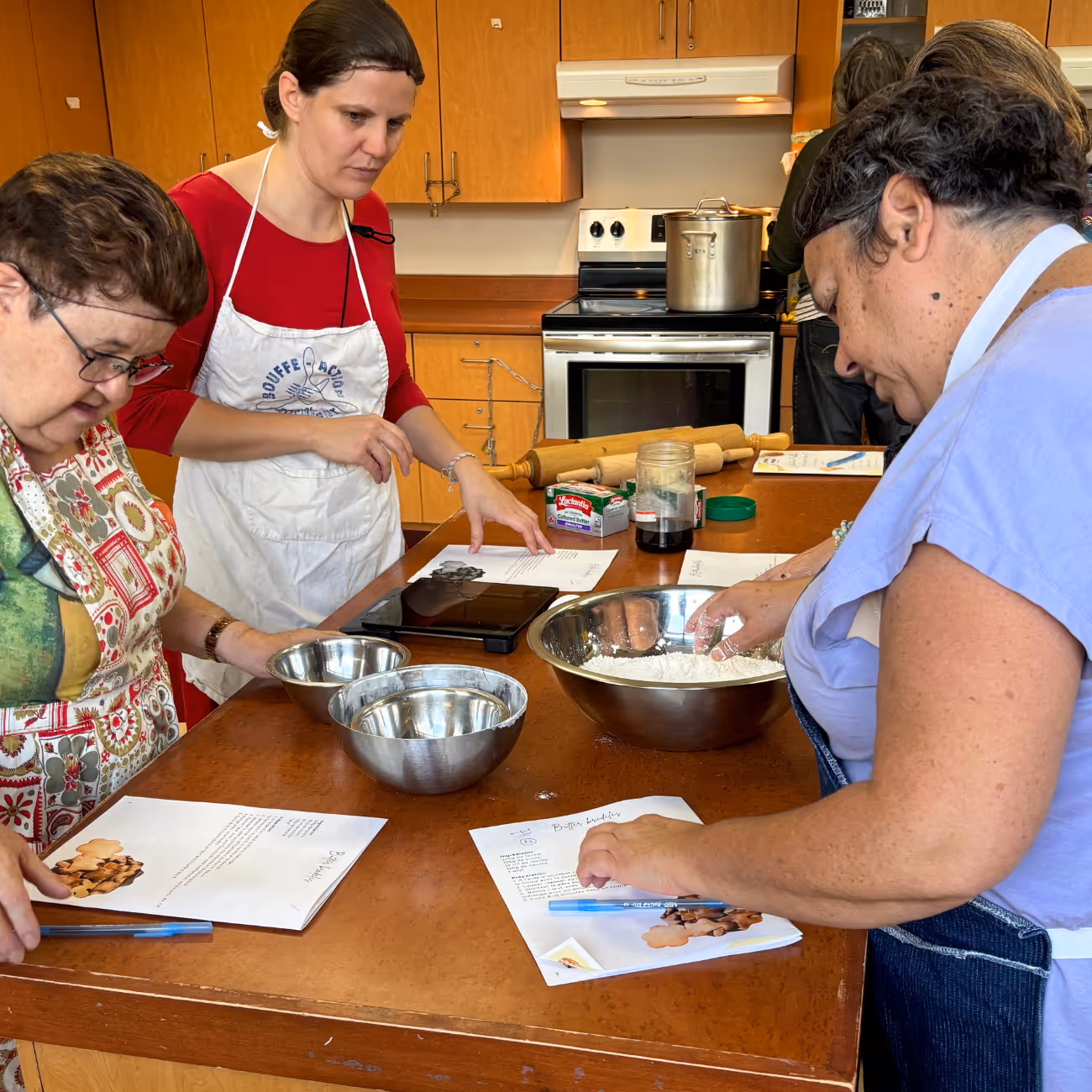 Quatre femmes dans une cuisine participent à un atelier de cuisine en préparant de la pâte avec des ingrédients et des recettes sur une table en bois.