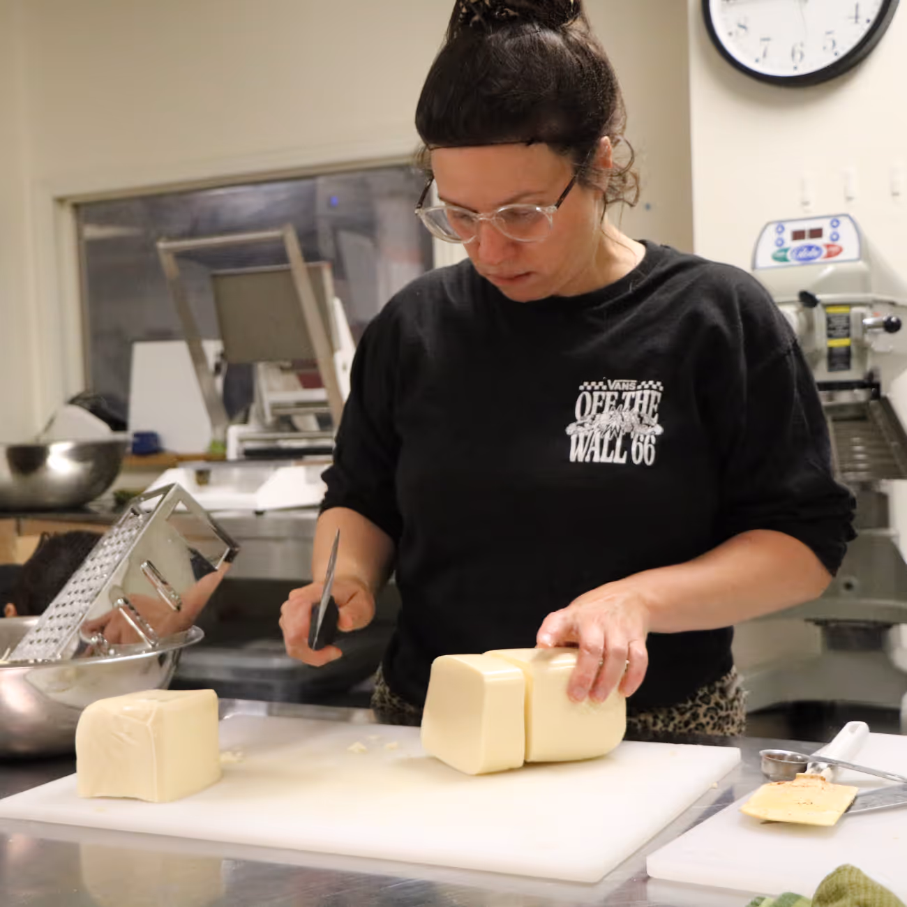 Une femme portant des lunettes coupe un grand bloc de fromage sur une planche blanche dans une cuisine industrielle.