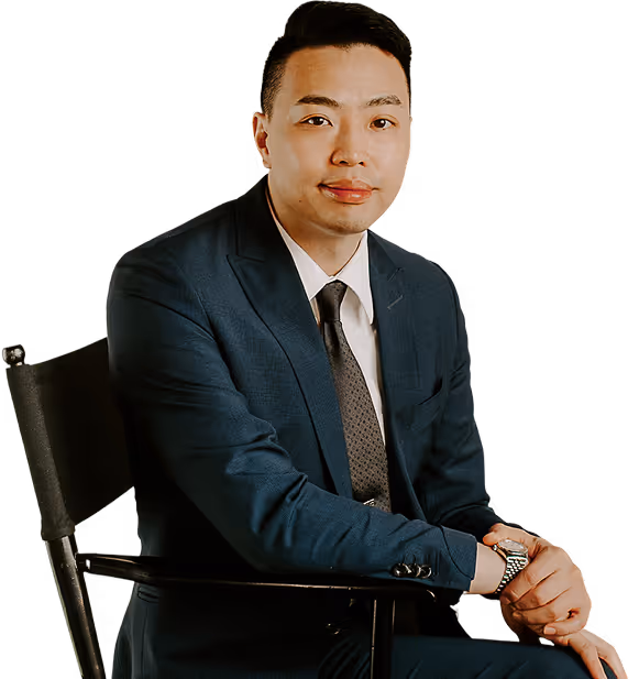 Man in a dark blue suit with a white shirt and tie sitting on a chair, hands clasped and smiling slightly.