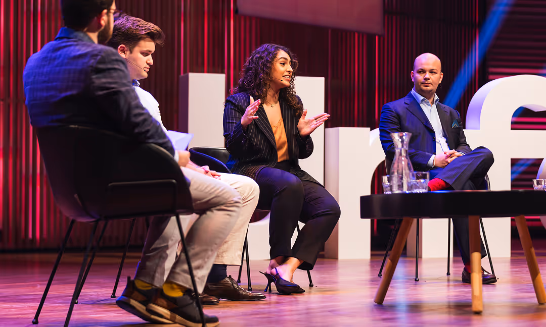 Four people sitting on chairs on a stage engaged in a panel discussion with a wooden table holding glassware in front of them.