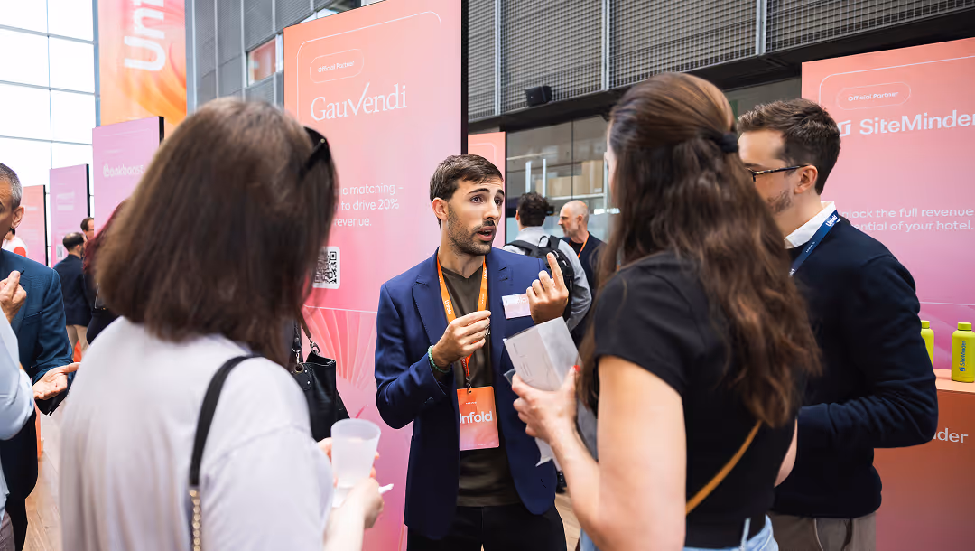 Man in a blazer explaining with hand gestures to a small group at a conference with pink promotional banners in the background.
