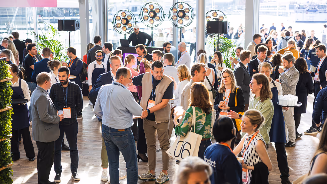 A busy indoor networking event with diverse people talking, shaking hands, and wearing conference badges.