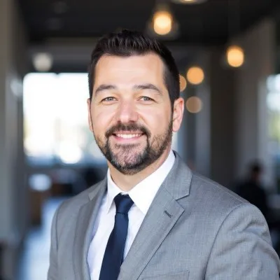 Smiling man with dark hair and beard wearing a gray suit and dark tie in a blurred indoor setting with hanging lights.