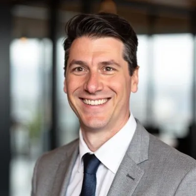 Smiling man with dark hair wearing a gray suit and navy tie in a blurred indoor setting.