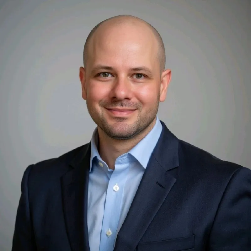 Smiling bald man wearing a dark suit jacket and light blue shirt against a gray background.