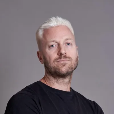Portrait of a man with short white hair and beard wearing a black shirt against a plain gray background.