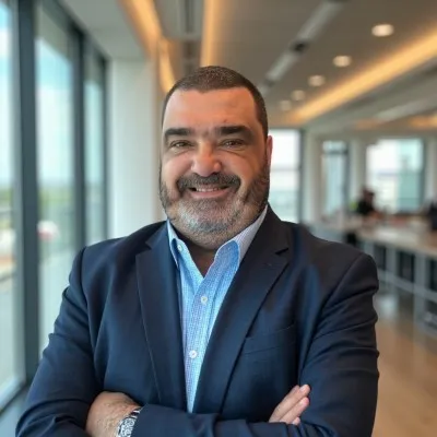 Smiling man in business attire with arms crossed standing by large windows in a modern office.