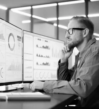 Man with glasses analyzing multiple data charts and graphs displayed on dual computer monitors in an office setting.