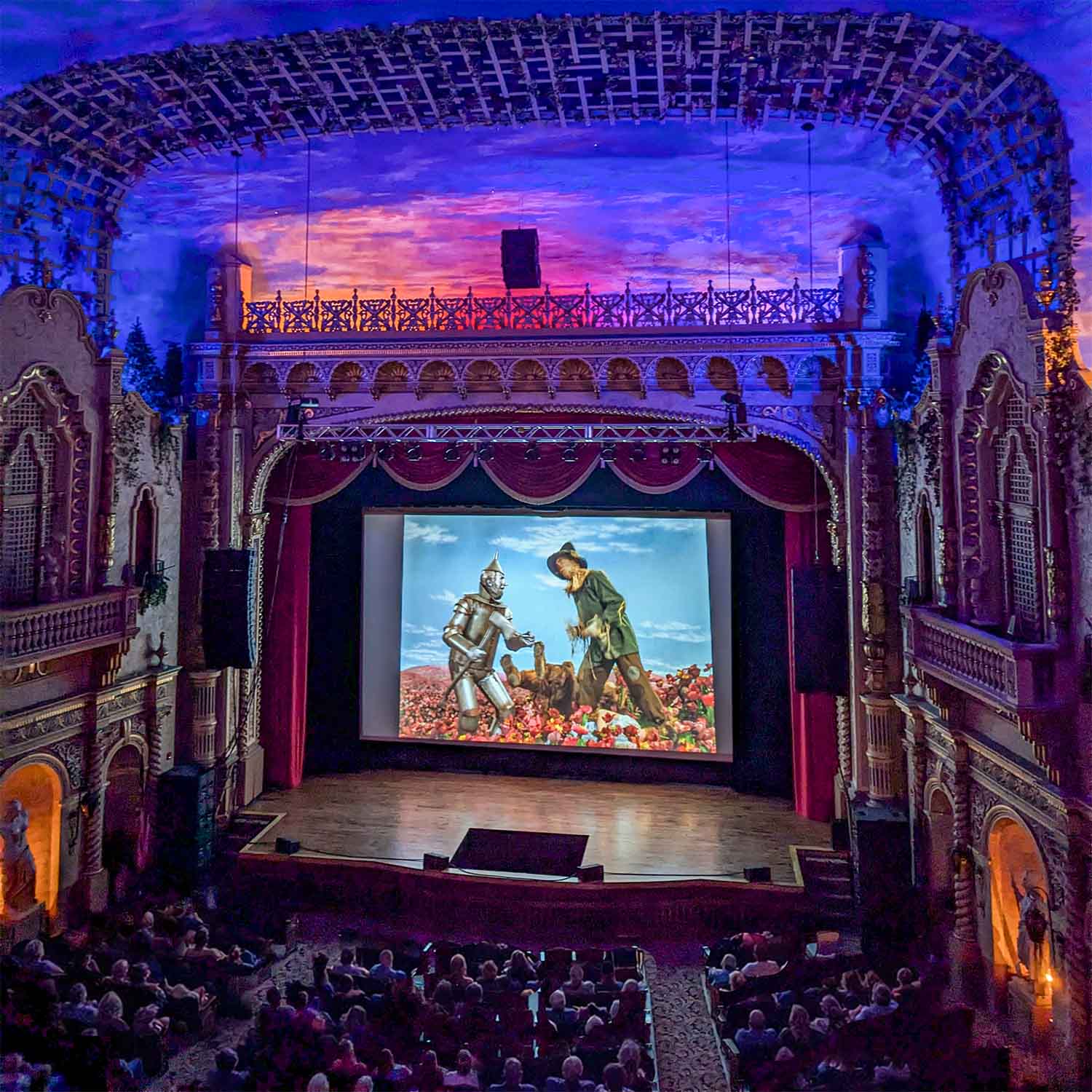 The Paramount Theatre interior with a large screen displaying a scene from The Wizard of Oz featuring the Tin Man and Scarecrow among flowers, with an audience watching.