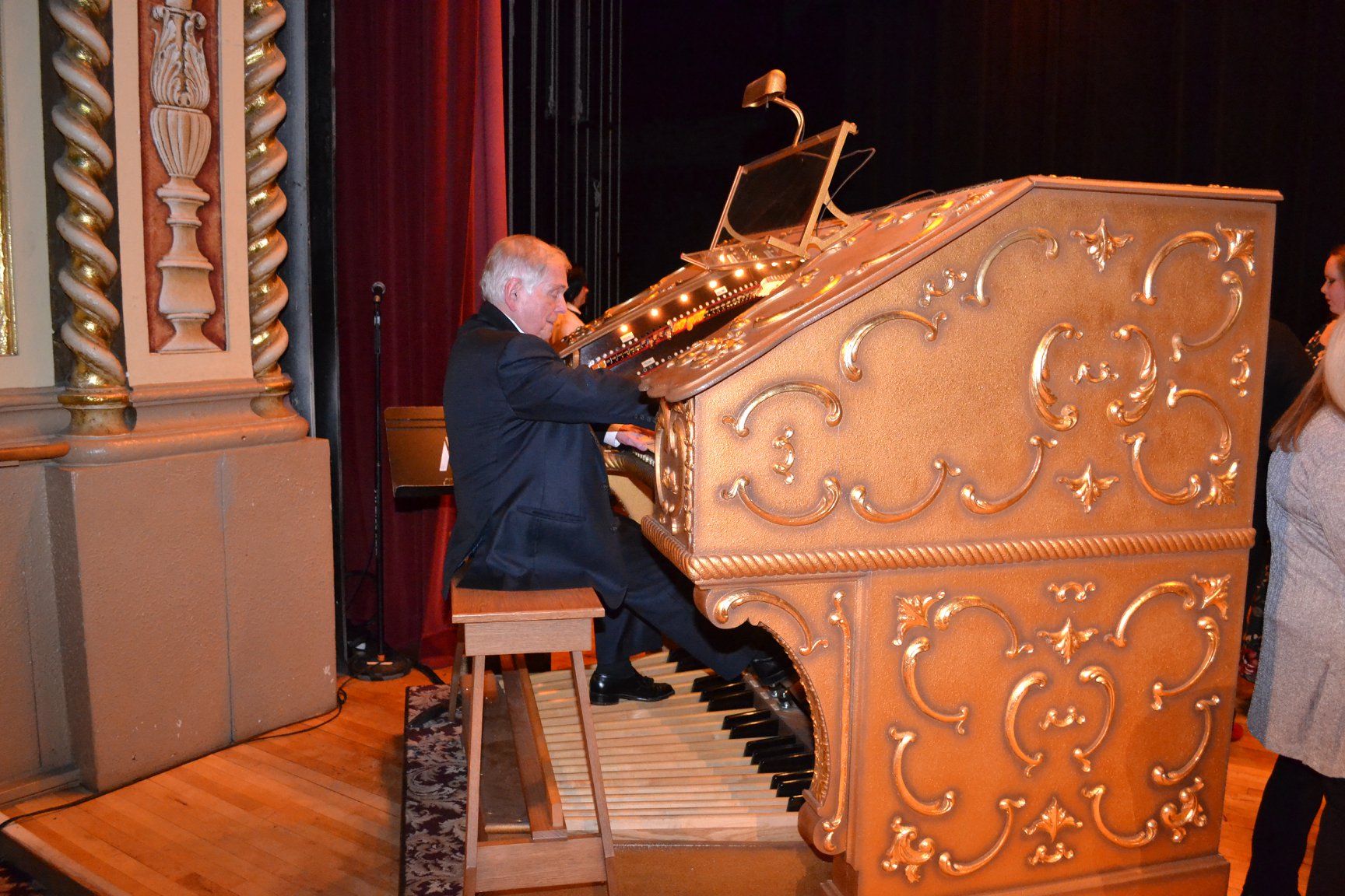 Man in formal attire playing a large ornate golden Page pipe organ on a stage.