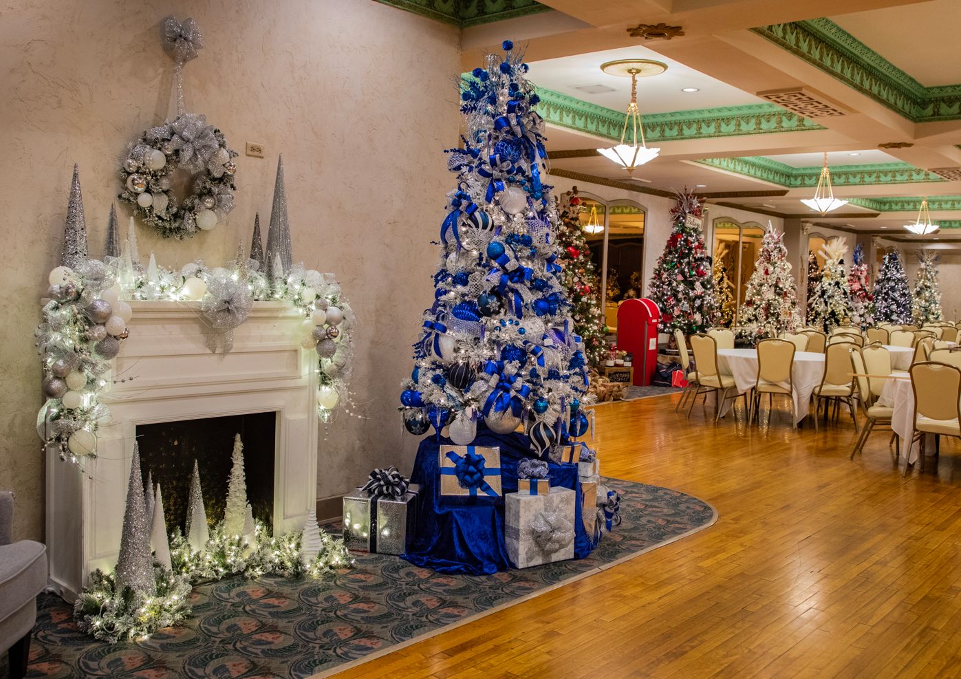 Indoor festive room with decorated blue and silver Christmas tree, white fireplace adorned with garland and ornaments, and several other decorated Christmas trees in the background at The Paramount Theatre.